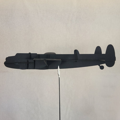 A black cardboard model of a British Lancaster bomber, a four engine military aircraft, side profile against a light gray textured background with a thin arrow shaft pointing horizontally to the right.