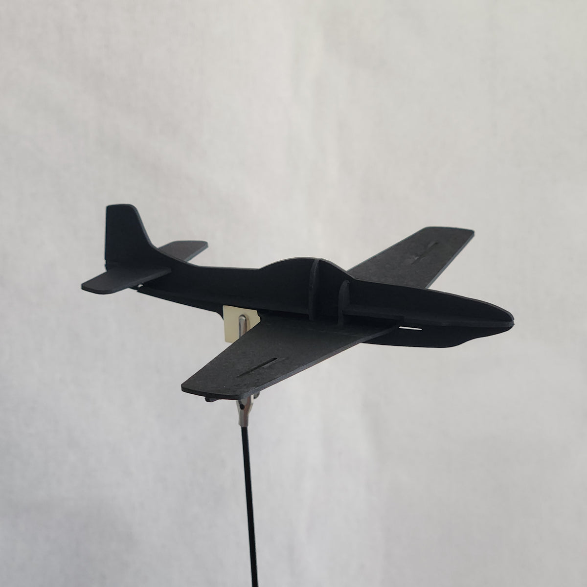 A black cardboard model of a U.S. Army P-51D Mustang airplane with a tail fin and a wire attached to a metal clip, shown against a plain light beige background.