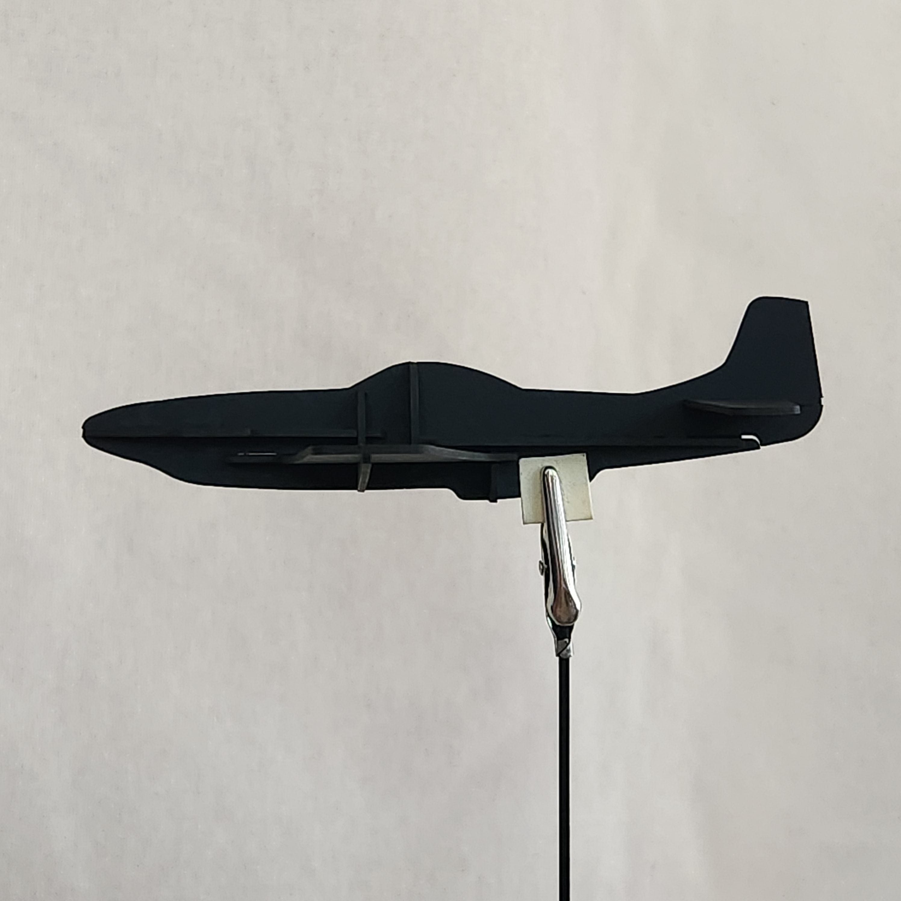 A black cardboard model of a U.S. Army P-51D Mustang airplane with a tail fin and a wire attached to a metal clip, shown against a plain light beige background.