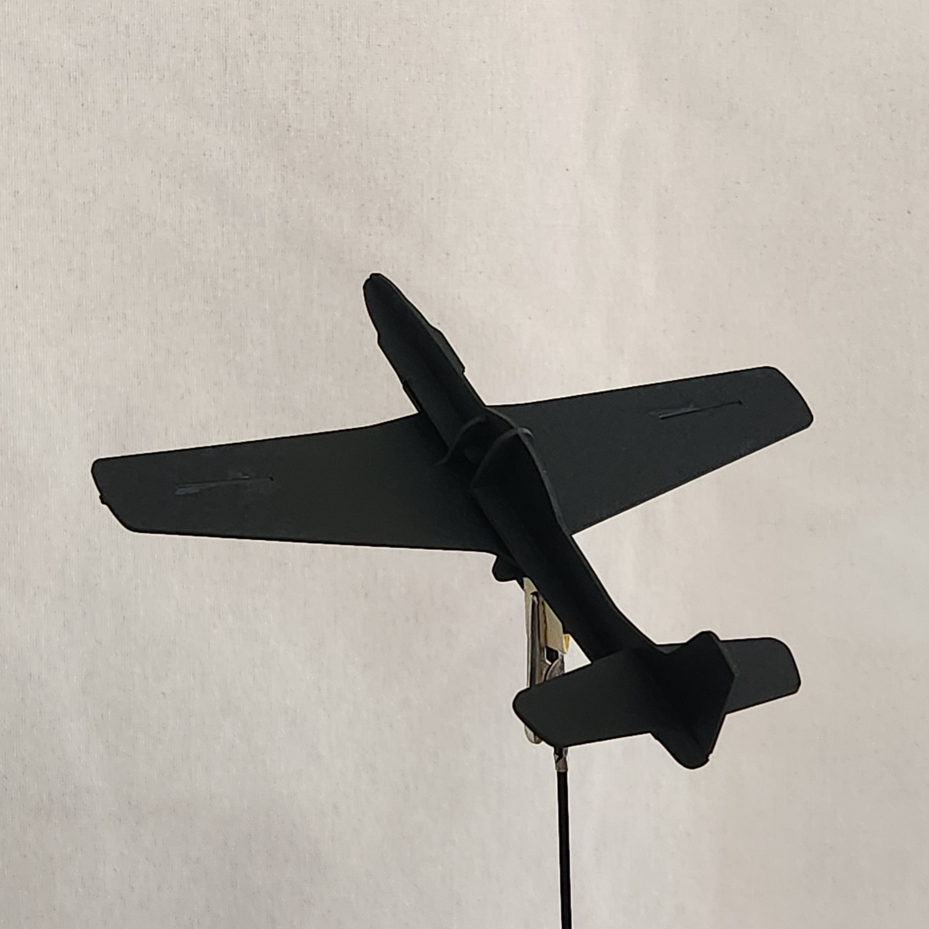 A black cardboard model of a U.S. Army P-51D Mustang airplane with a tail fin and a wire attached to a metal clip, shown against a plain light beige background.